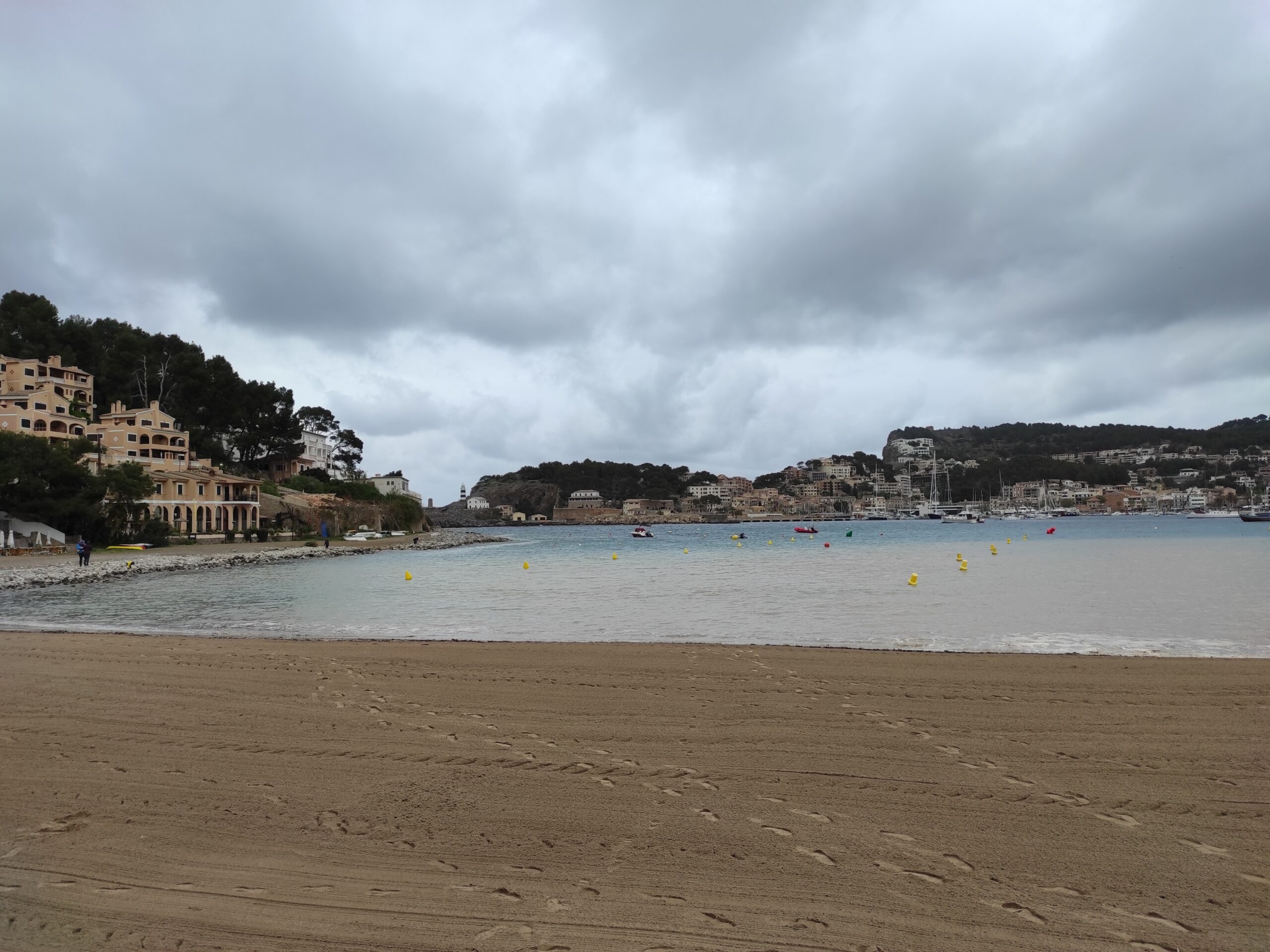 Port de Sóller Mallorca: twee stranden in een ronde baai 3 Het brede, vrijwel lege zand van Platja d'en Repic aan de westkant van Port de Sóller, met gele boeien in het vlakke water en aan de overkant de havenfront en de beboste kaap onder een zware grijze lucht