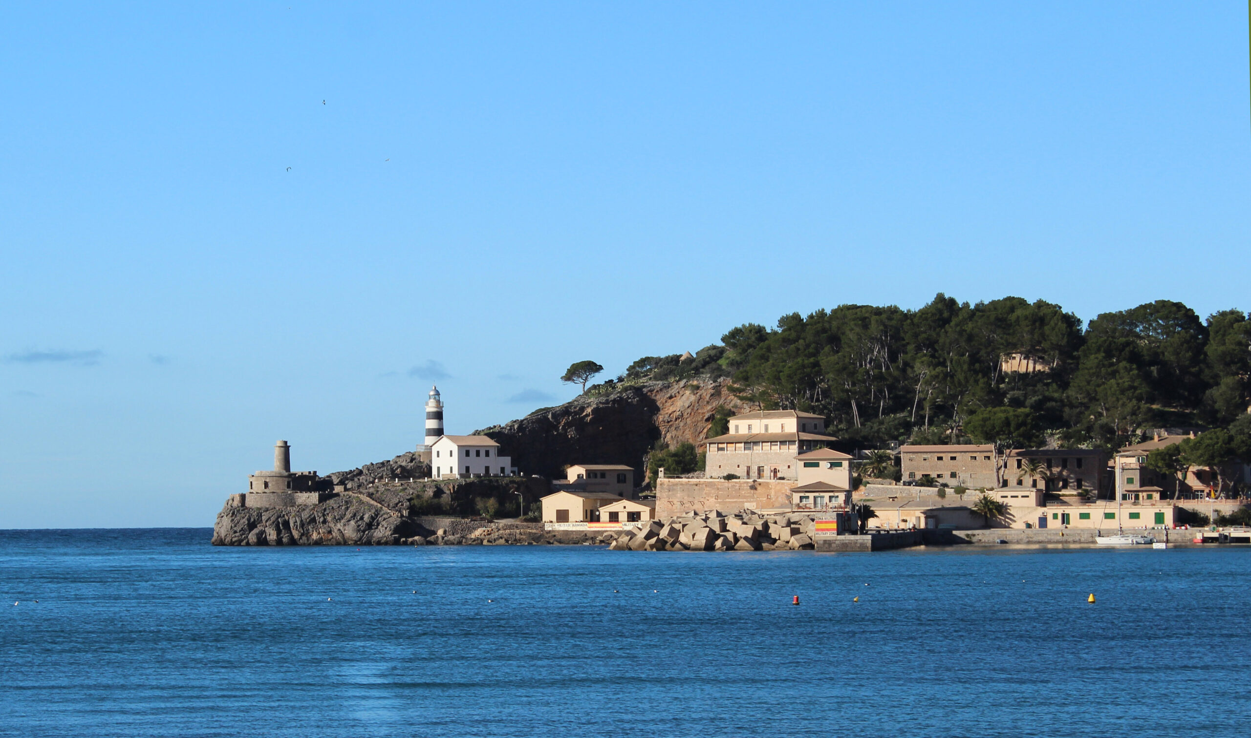 Port de Sóller Mallorca: twee stranden in een ronde baai 4 De rotspunt aan de monding van de baai van Port de Sóller met de zwart-wit gebande vuurtoren Far de sa Creu, de ronde stenen wachttoren ernaast en de lage gebouwen van de kade aan de waterlijn, gezien vanaf Platja d'en Repic