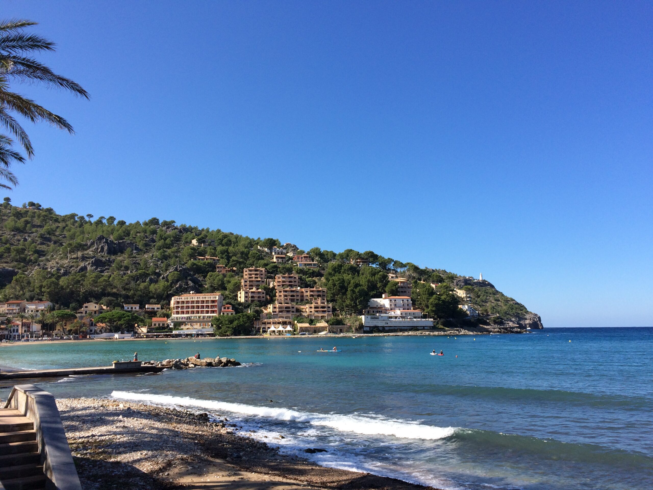 Port de Sóller Mallorca: twee stranden in een ronde baai 5 De kiezelrand van Platja des Través aan de oostkant van de baai van Port de Sóller, met een golf die op het grind breekt, een korte stenen strekdam en aan de overkant de beboste helling met appartementen die doorloopt tot aan de kaap