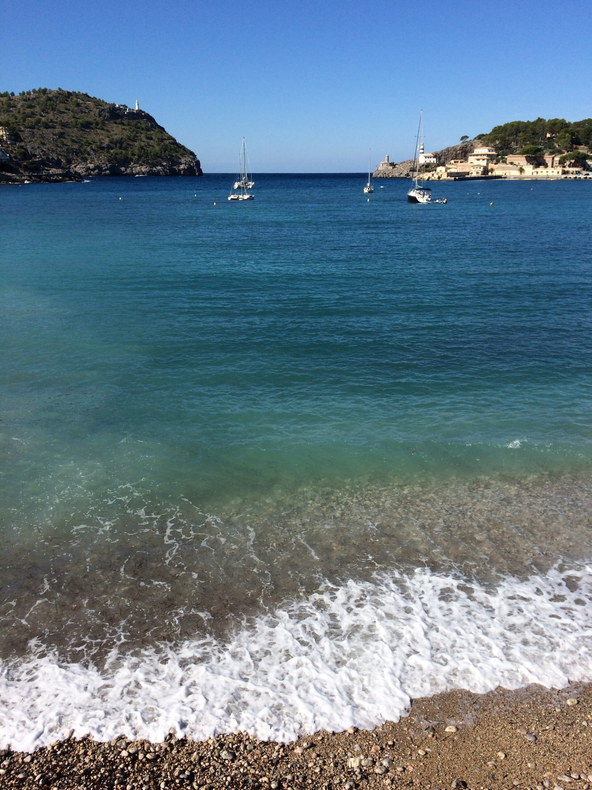 Port de Sóller Mallorca: twee stranden in een ronde baai 6 Helder water over de kiezels aan Platja des Través, met zeilboten voor anker in de baai van Port de Sóller en de twee landpunten die de monding bijna sluiten, rechts die met de vuurtoren