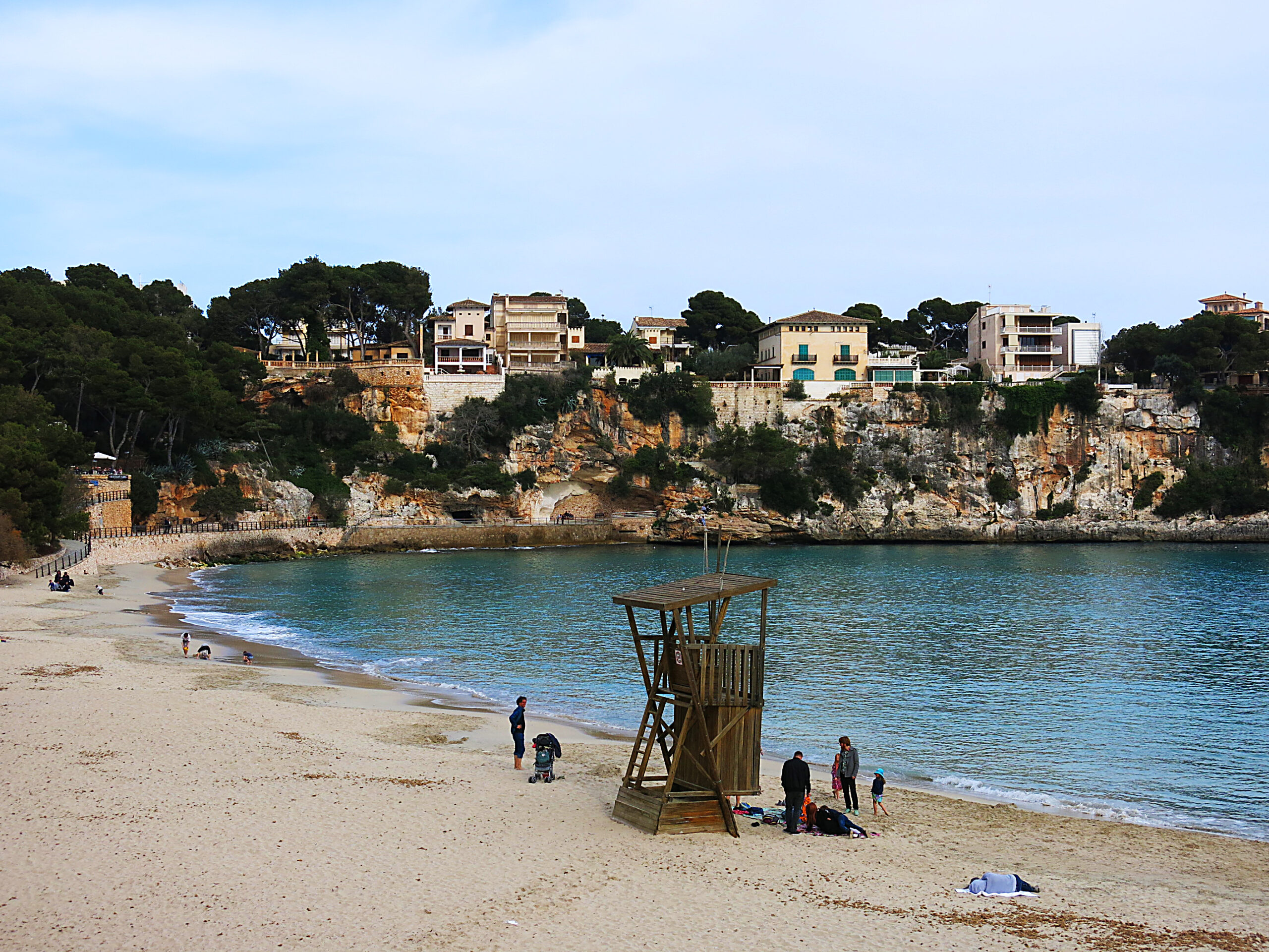 Porto Cristo Mallorca: strand, grotten en de landing van 1936 3 Het rustige strand van Porto Cristo buiten het seizoen, met een houten badmeesterspost op het zand en de okerkleurige kliffen van ses Coves Blanques met hun huizen erachter