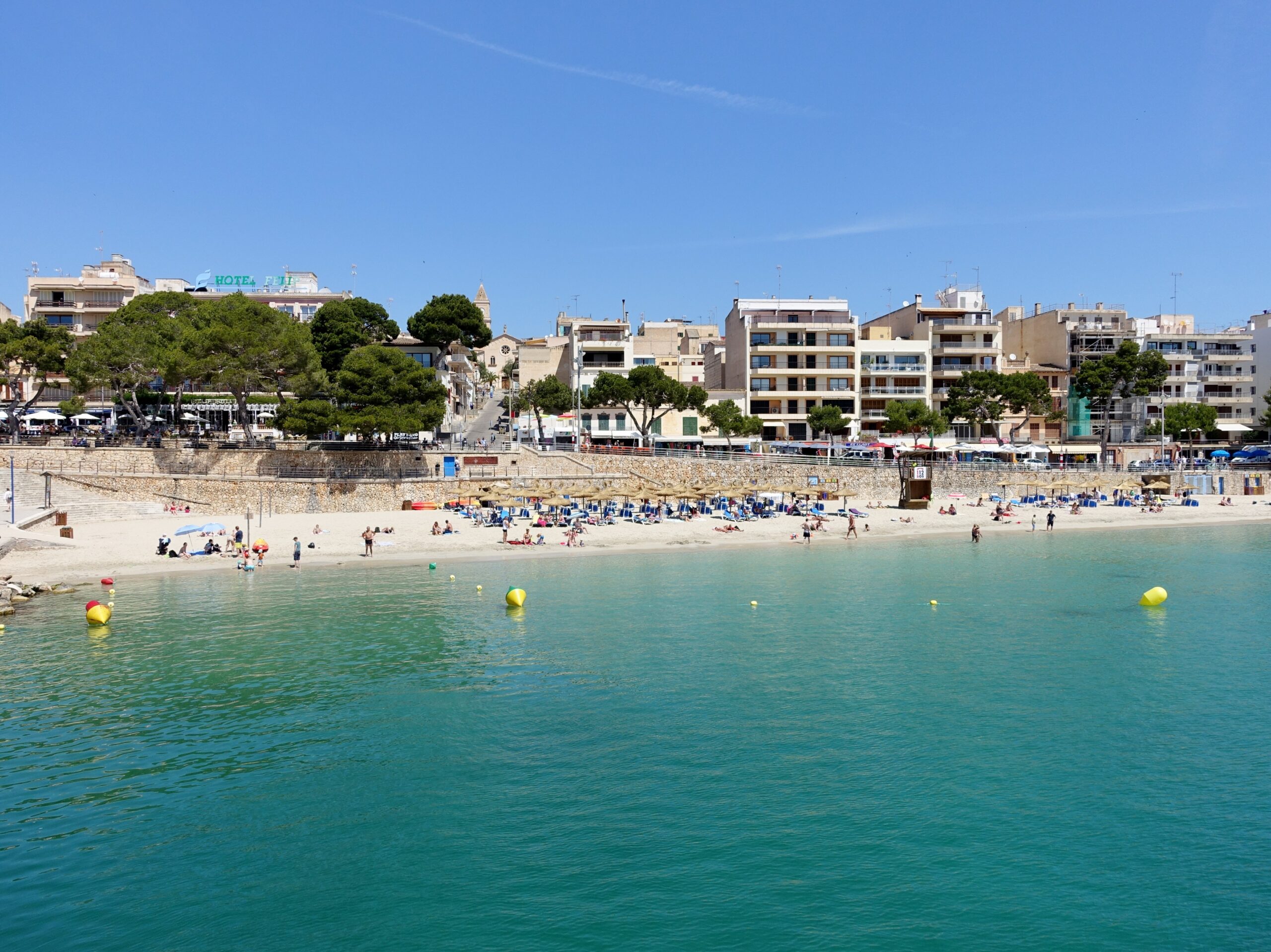 Porto Cristo Mallorca: strand, grotten en de landing van 1936 4 Het strand van Porto Cristo in juni, gezien over het turquoise water van de inham, met rijen ligbedden op het zand en de huizen van het dorp erboven