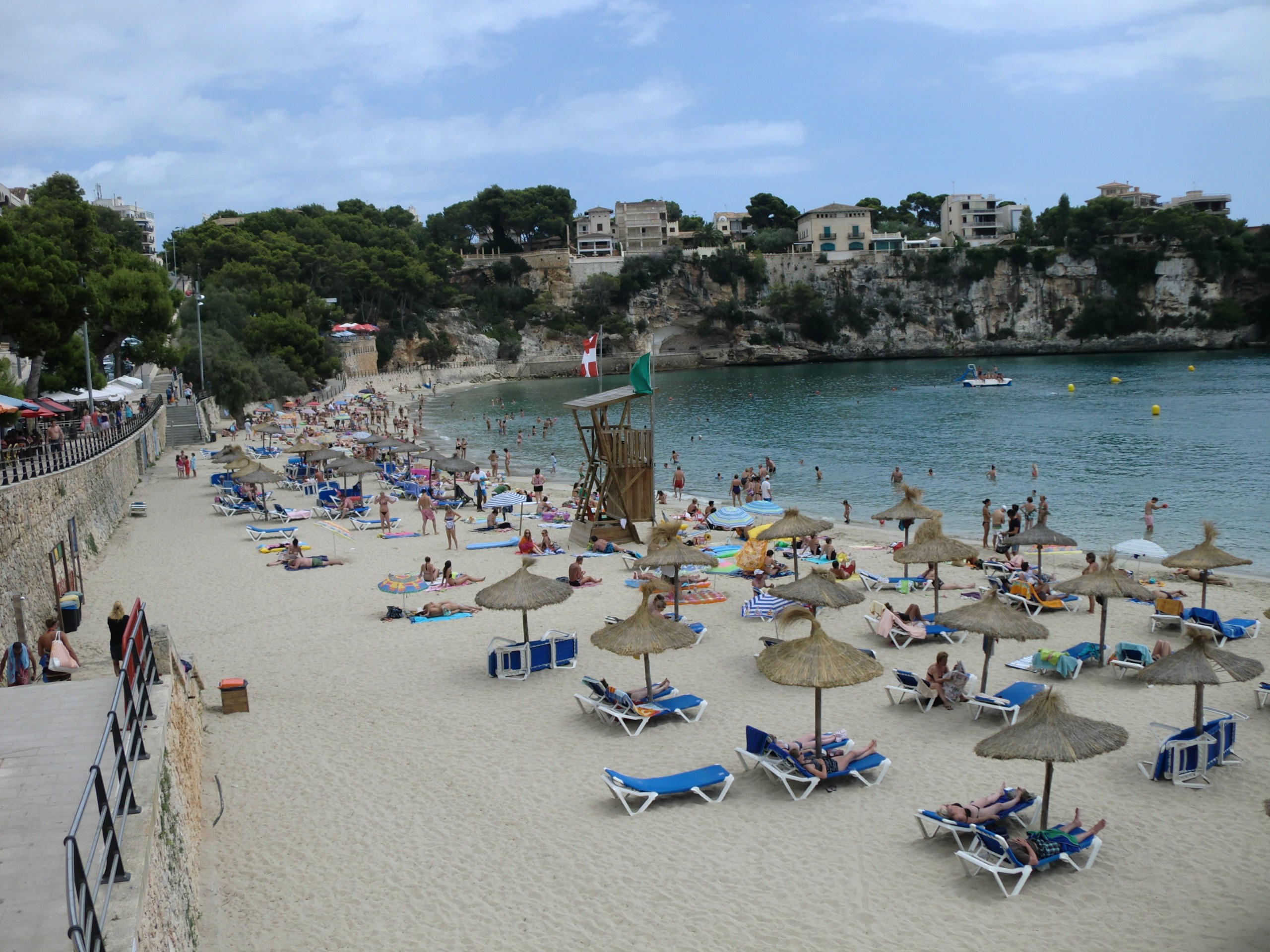 Porto Cristo Mallorca: strand, grotten en de landing van 1936 5 Strohutparasols en blauwe ligbedden op het zand van Porto Cristo, met links de gemetselde promenade en aan de overkant de begroeide klif