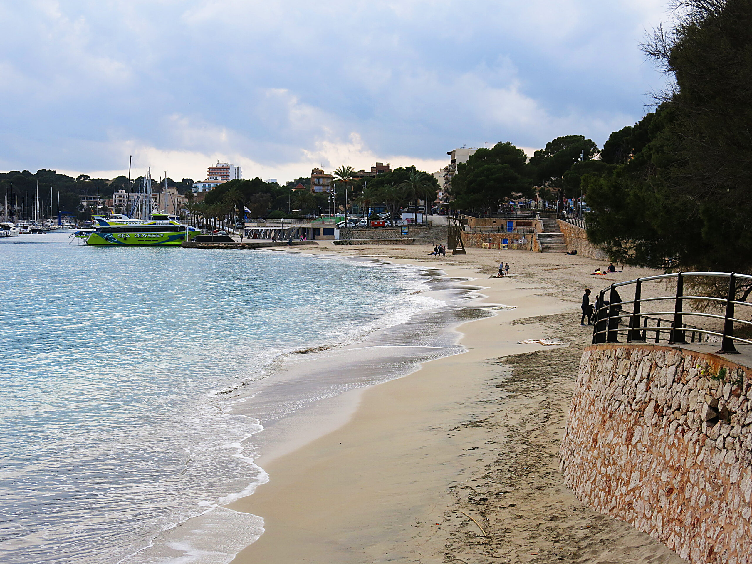 Porto Cristo Mallorca: strand, grotten en de landing van 1936 7 De boog van het strand van Porto Cristo bij bewolkt weer, met de gemetselde promenade rechts en de veerboot aan de kade in de verte