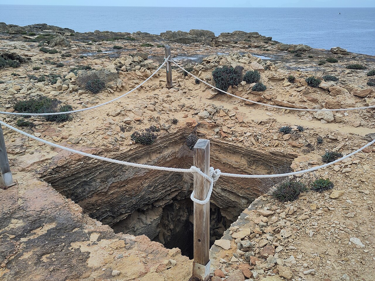 Met een touw afgezette opening in het rotsplateau bij Punta de sa Pedrera, met de zee erachter