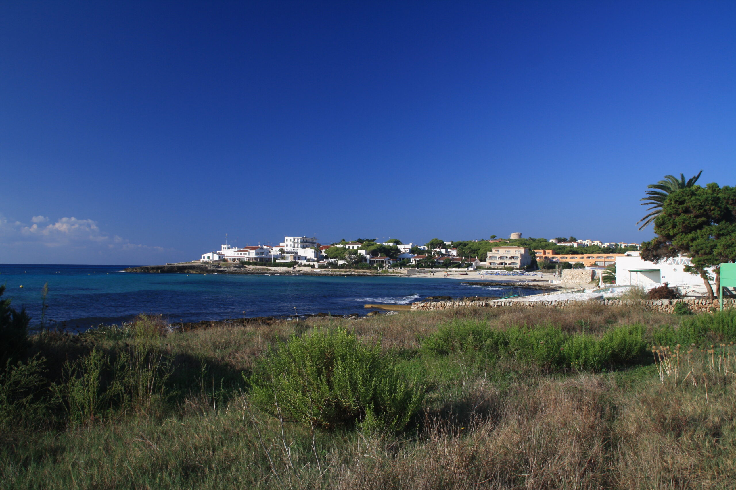 De baai van Punta Prima op Menorca met witte appartementen, het strand met parasols en de Torre de Son Ganxo op de heuvel