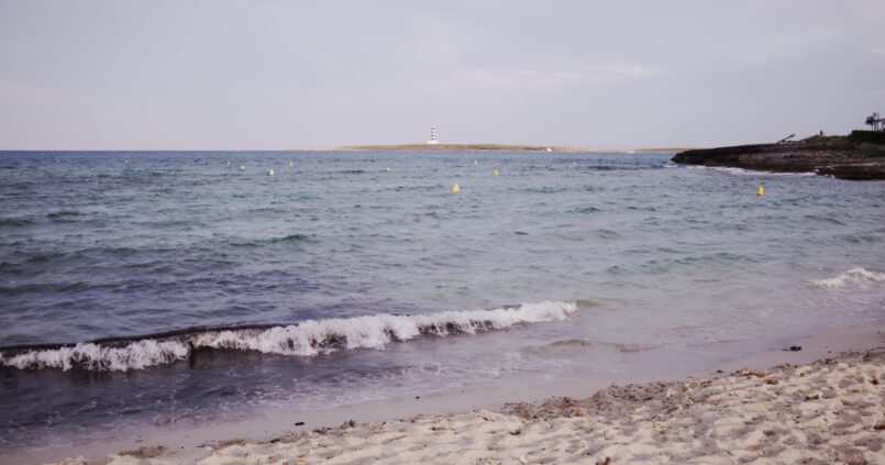 Het witte zand van Punta Prima op Menorca met het eilandje Illa de l'Aire en zijn gestreepte vuurtoren aan de horizon
