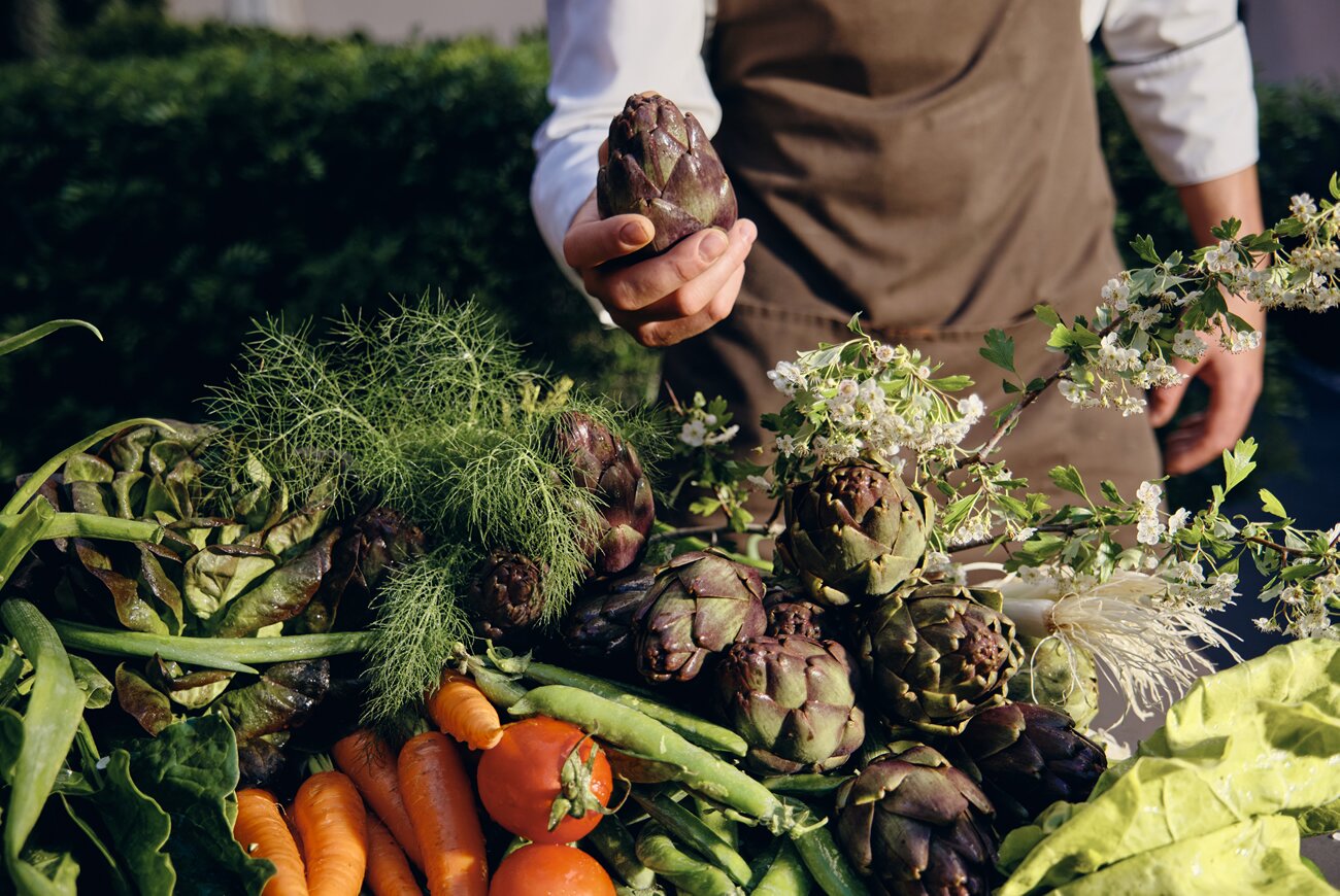 Quadrat in Palma: eten in de tuin van Hotel Sant Francesc 7 Handen van een kok boven een stapel verse groenten met artisjokken, wortels, tomaten en venkel van Mallorcaanse telers
