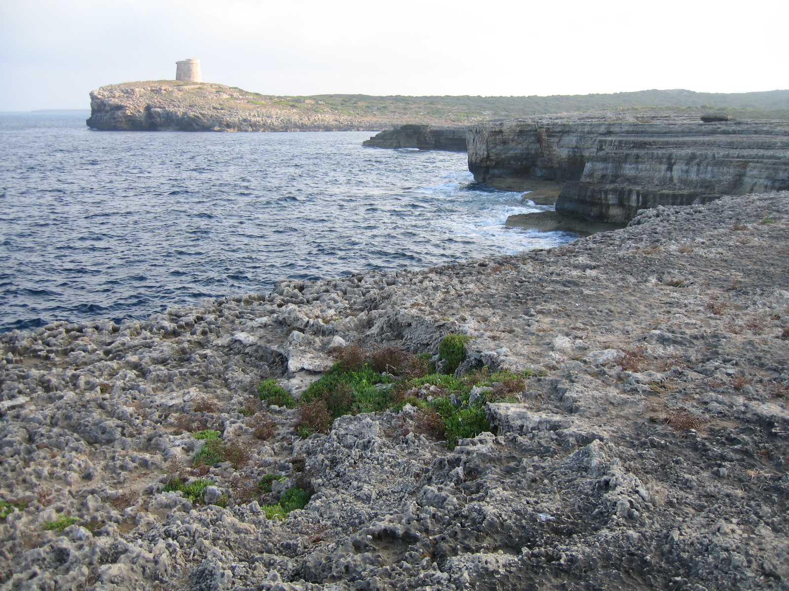 De lage rotskust van de zuidoosthoek van Menorca met een wachttoren op de kaap in de verte