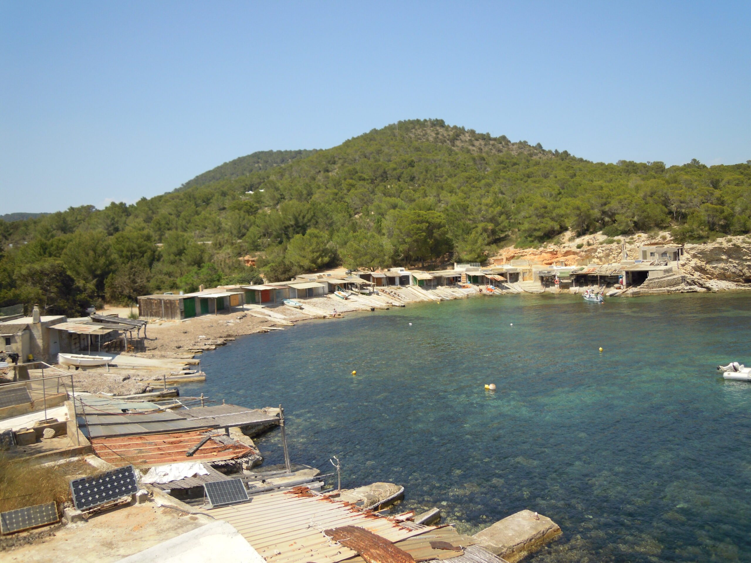 Het vissershaventje van Sa Caleta met de rij bootloodsen met gekleurde deuren, de betonnen hellingen naar het water en de beboste Puig d'en Toni Fita erachter
