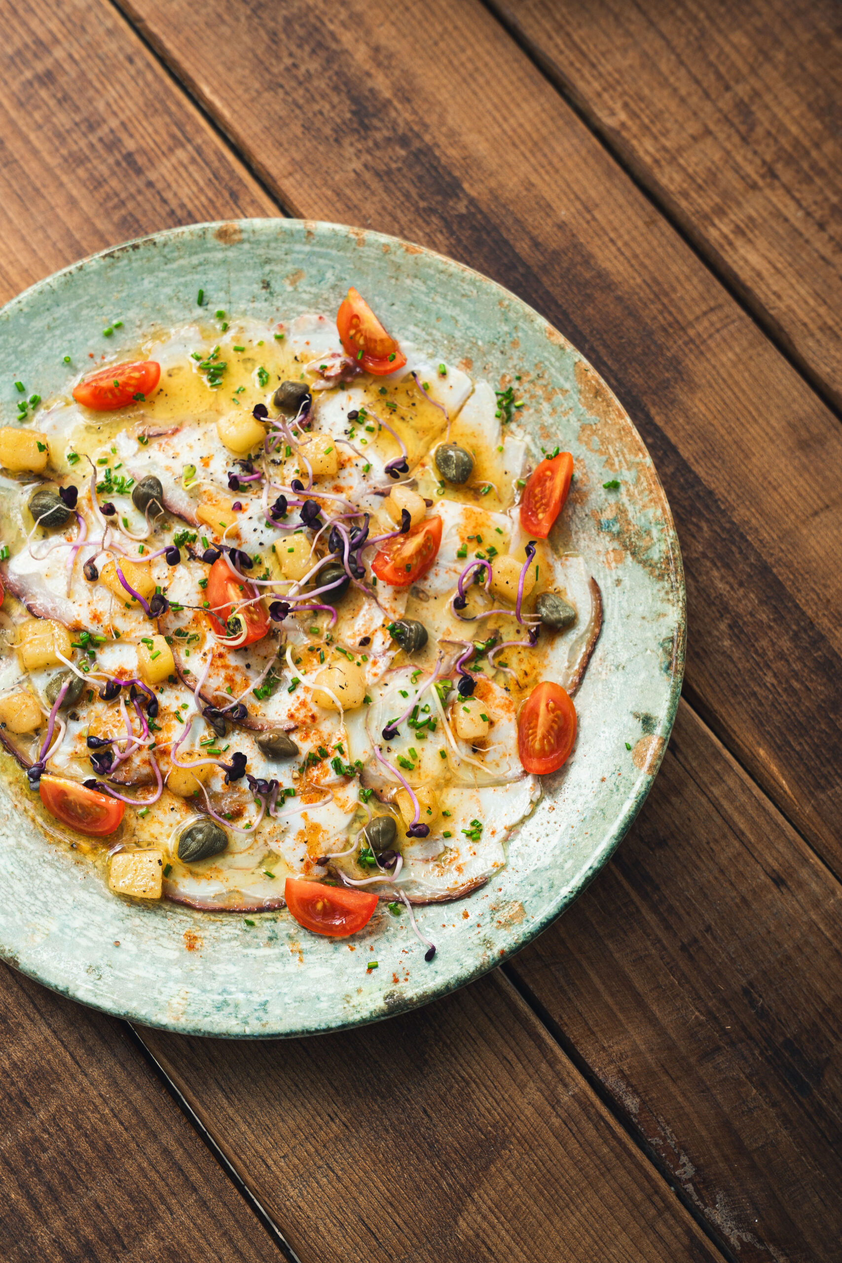 Carpaccio van octopus op een groen geglazuurd bord met blokjes aardappel, cherrytomaat, kappertjes, bieslook en gerookte paprikapoeder