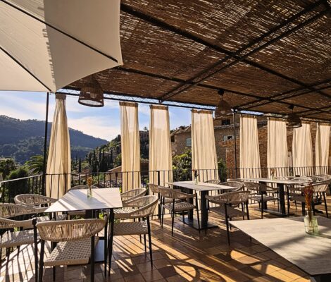 Het overdekte terras van Sa Font Fresca in Deià met rieten dak, witte gordijnen en gevlochten stoelen, en uitzicht op de beboste bergen en de stenen huizen van het dorp