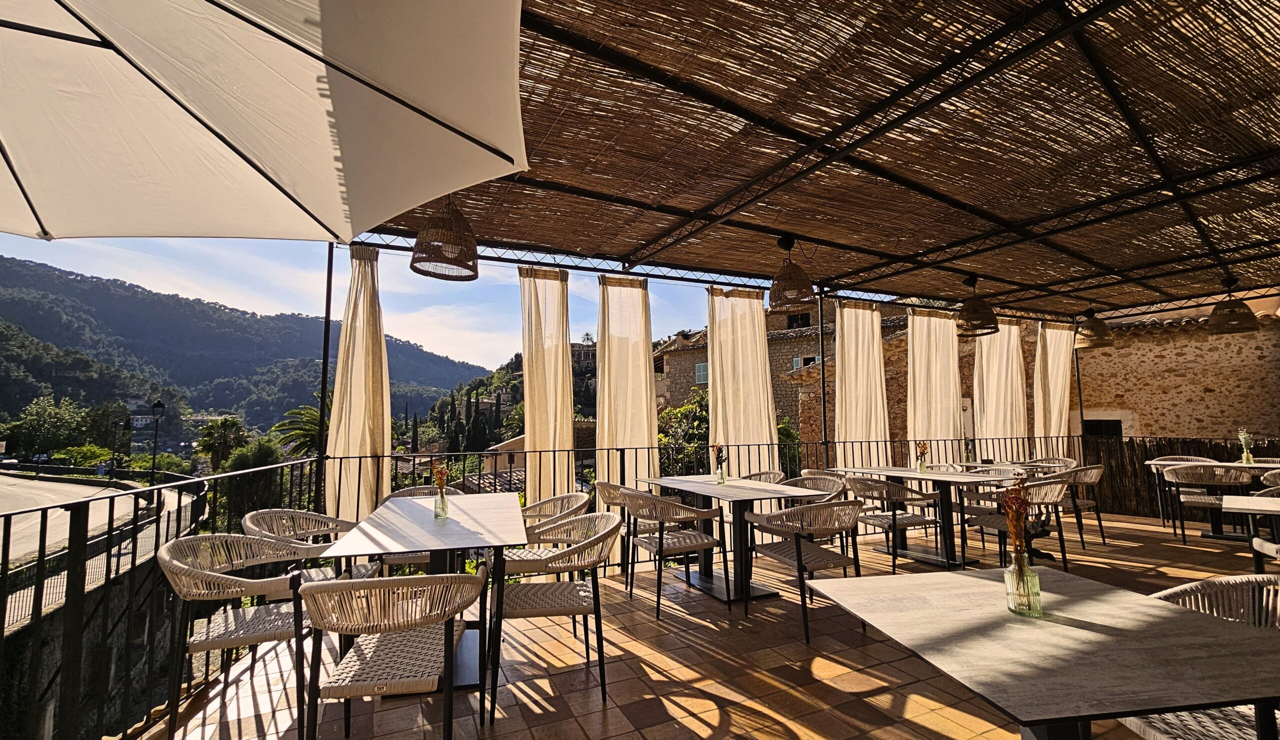 Het overdekte terras van Sa Font Fresca in Deià met rieten dak, witte gordijnen en gevlochten stoelen, en uitzicht op de beboste bergen en de stenen huizen van het dorp