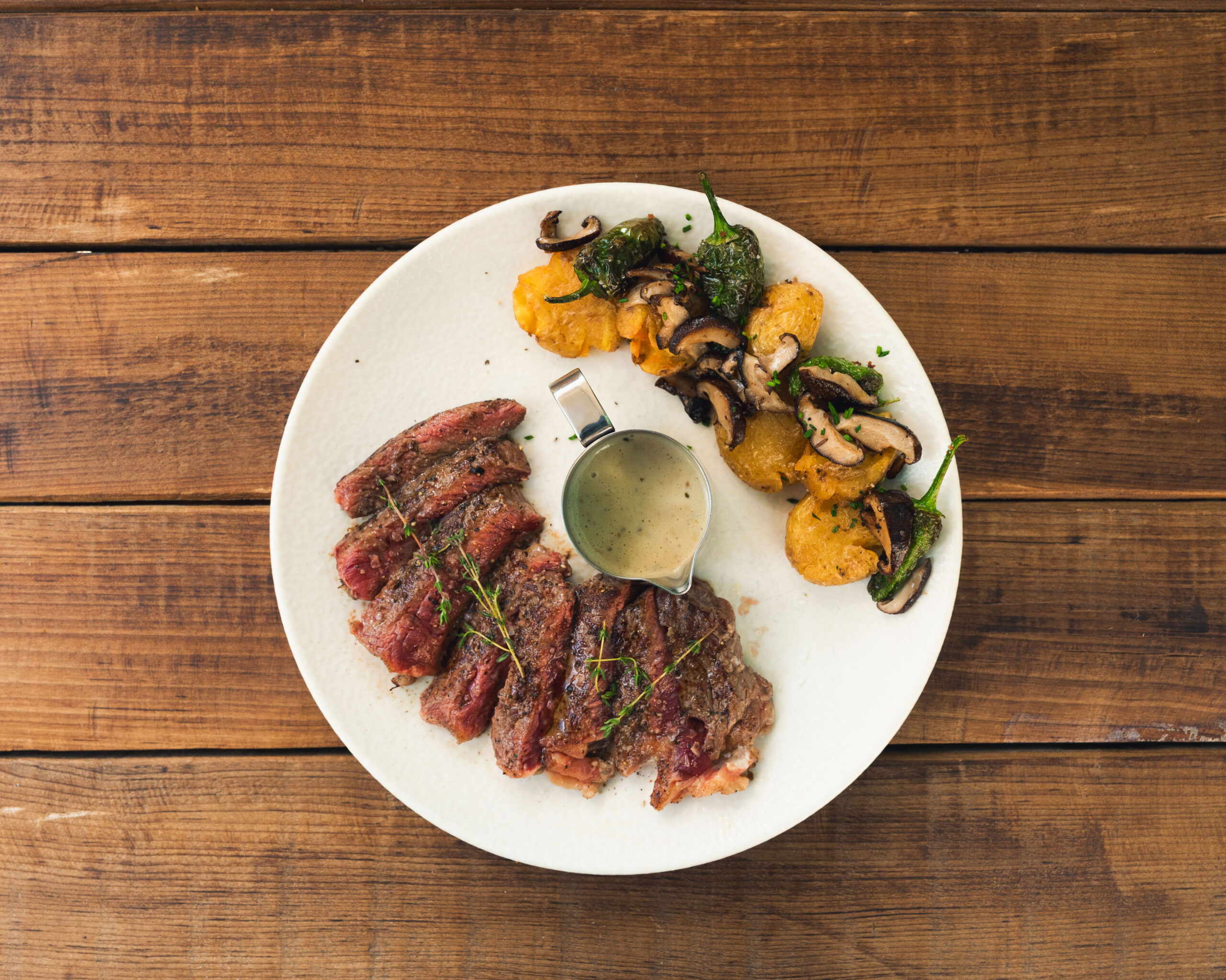 Tagliata van Angus-entrecote in plakken op een wit bord, met gebakken aardappel, shiitake, padrónpepertjes en een kannetje saus, op een houten tafel