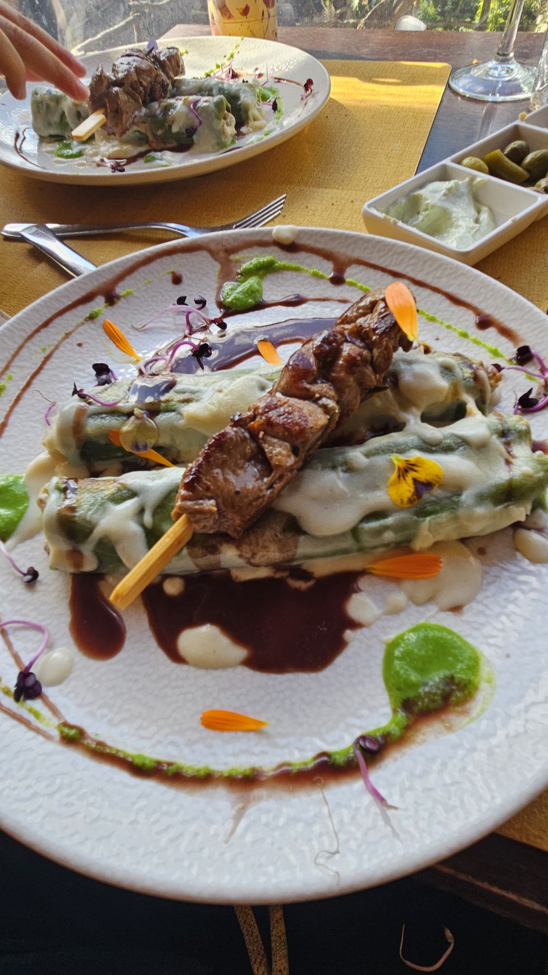 Canelones met groene pastavellen en bechamelsaus, met een lamsspies erop, afgewerkt met eetbare bloemen op een wit bord