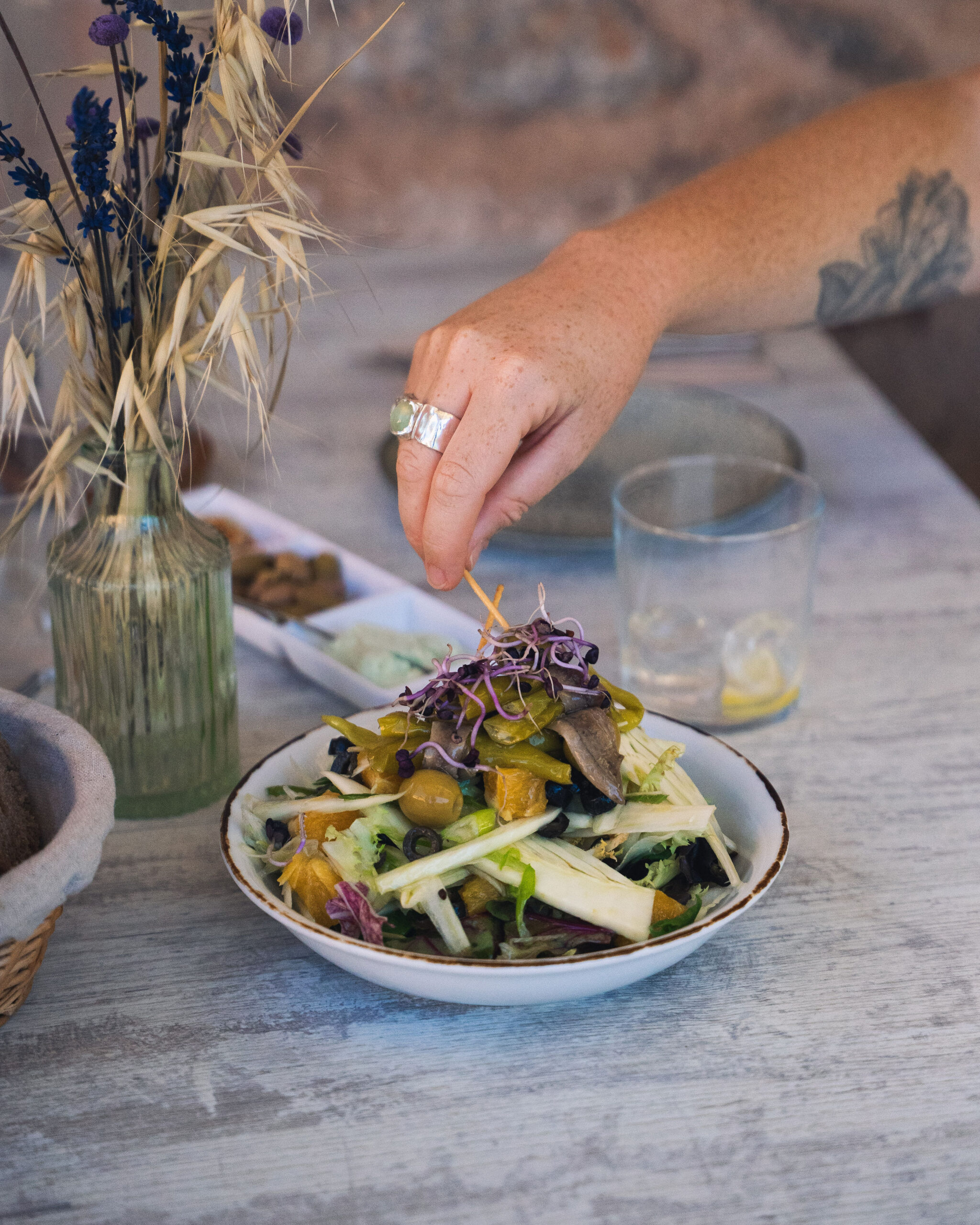 Een hand zet de laatste garnering op een salade met groene peper, olijven en sinaasappel, naast een broodmandje en een vaasje droogbloemen