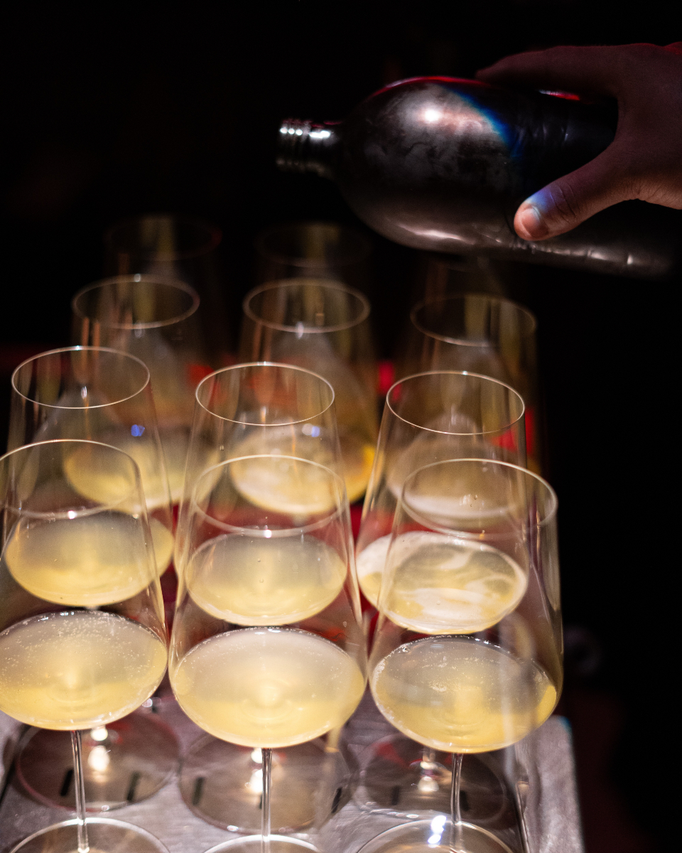 Een hand schenkt uit een donkere fles in een rij gevulde glazen op een dienblad in het donkere interieur van Sala de Personal in Palma