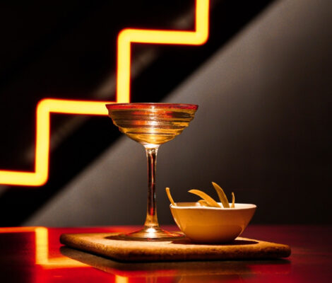Sala de Personal Palma: de cocktailkelder onder Ginbo (in pauze) 5 Een gouden cocktail in een coupe naast een schaaltje met een hapje, op een houten plankje voor de oranje neonbuis in de vorm van een trap bij Sala de Personal in Palma