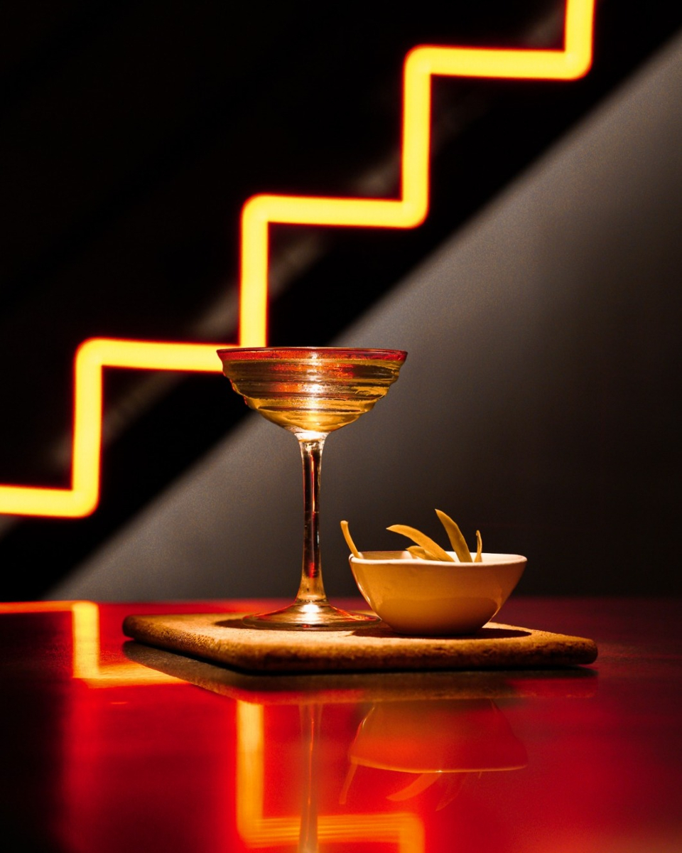 Een gouden cocktail in een coupe naast een schaaltje met een hapje, op een houten plankje voor de oranje neonbuis in de vorm van een trap bij Sala de Personal in Palma