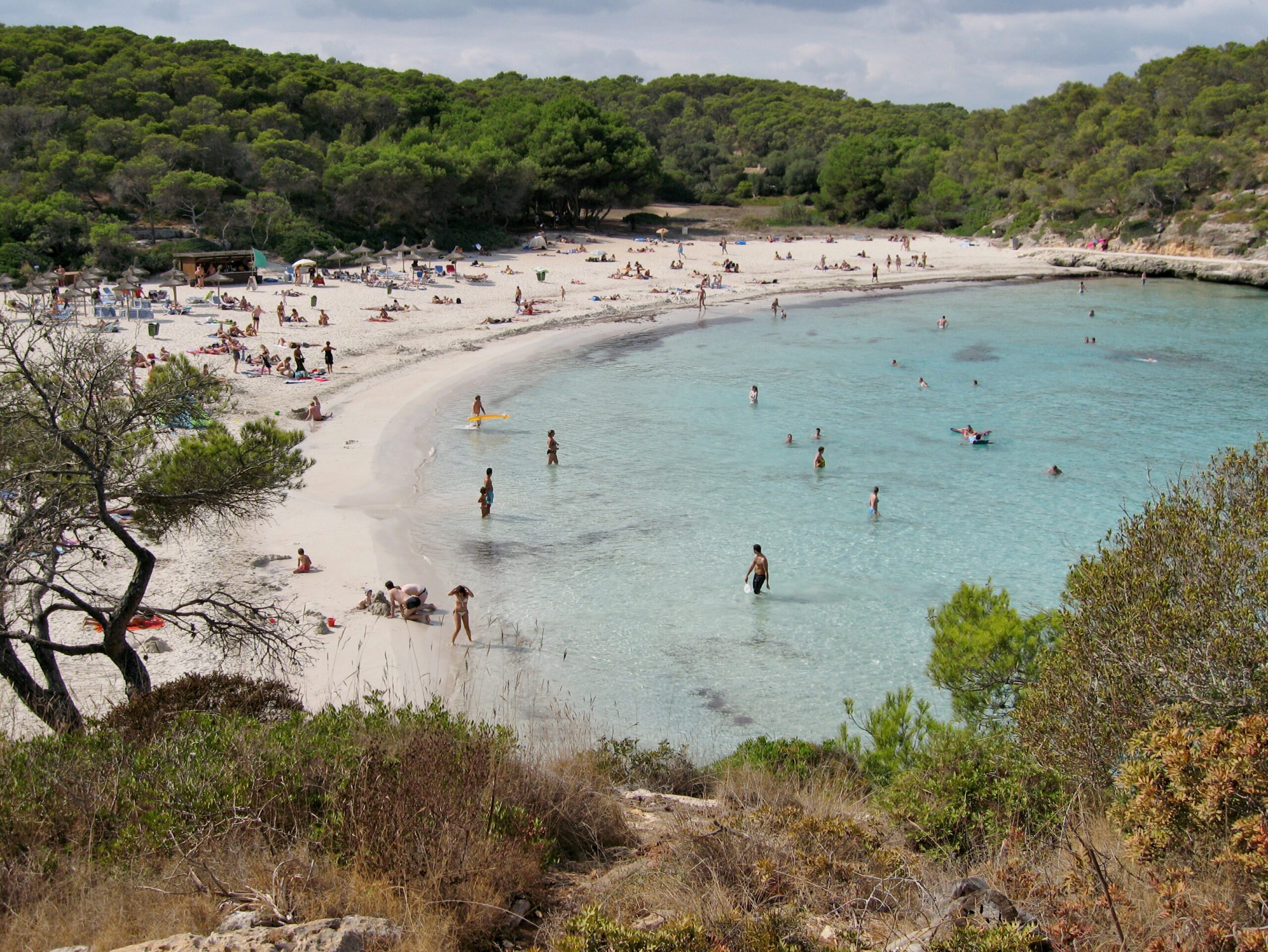 De boog van S'Amarador gezien vanaf het pad door het pijnbos, met badgasten in het ondiepe turquoise water