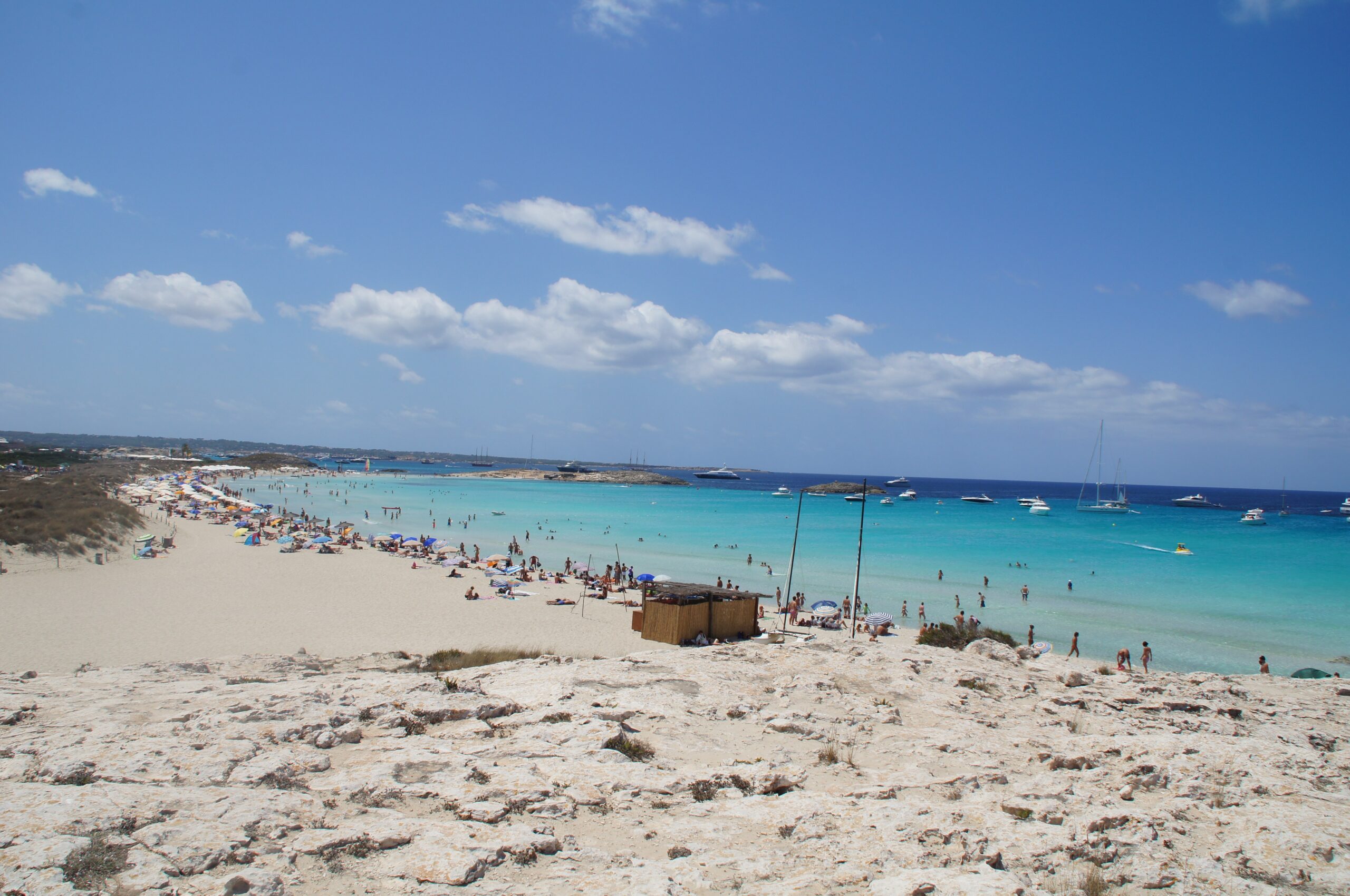 Ses Illetes Formentera: strandgids met de regels van 2026 2 Het witte zand van Ses Illetes in de zomer, met parasols langs de waterlijn, turquoise ondiep water en jachten voor de kust