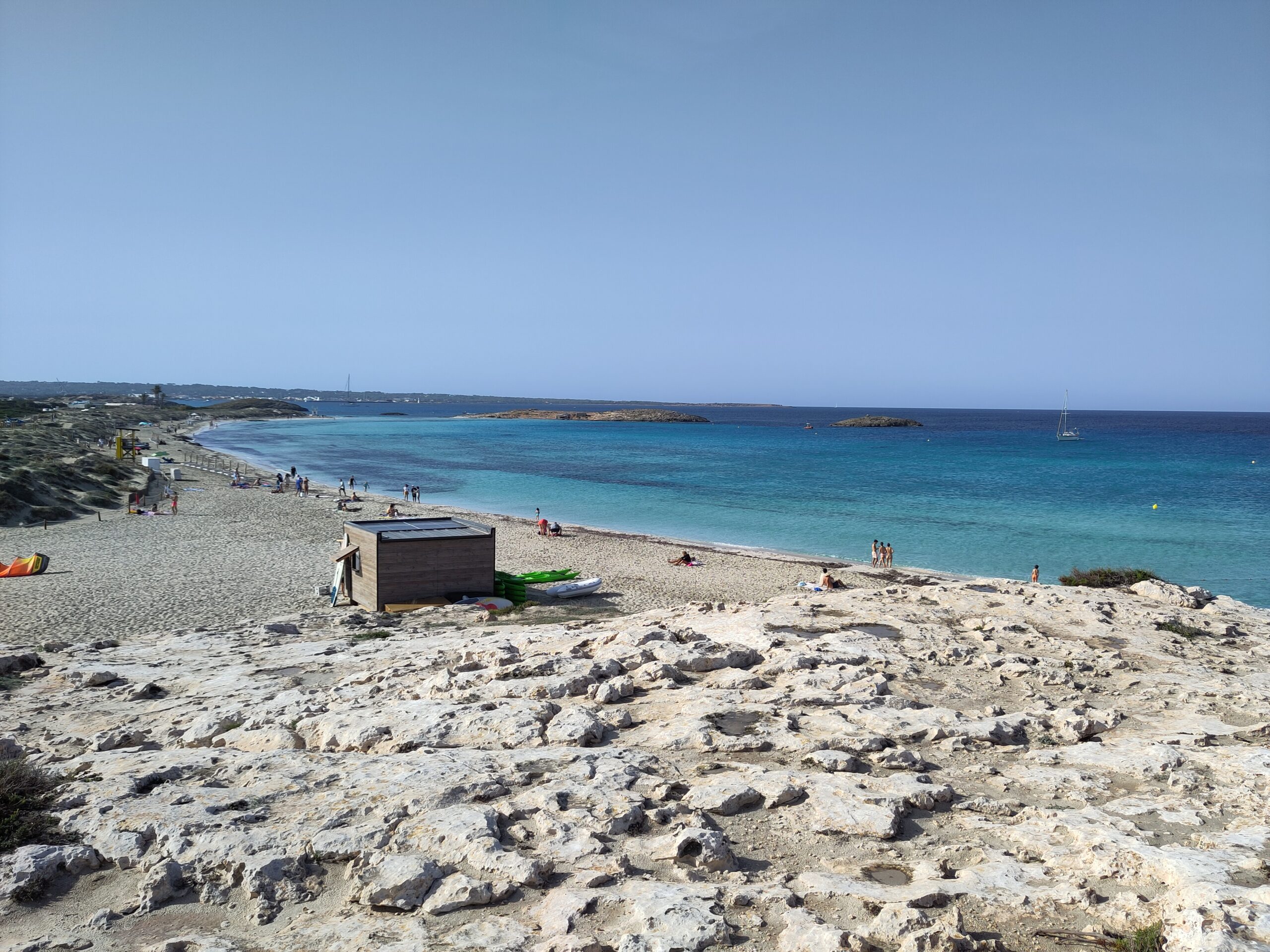 Ses Illetes Formentera: strandgids met de regels van 2026 4 Platja de n'Adolf in het najaar, met de lage rotspartij die het strand in tweeen deelt, een houten kajakhut en de eilandjes voor de kust
