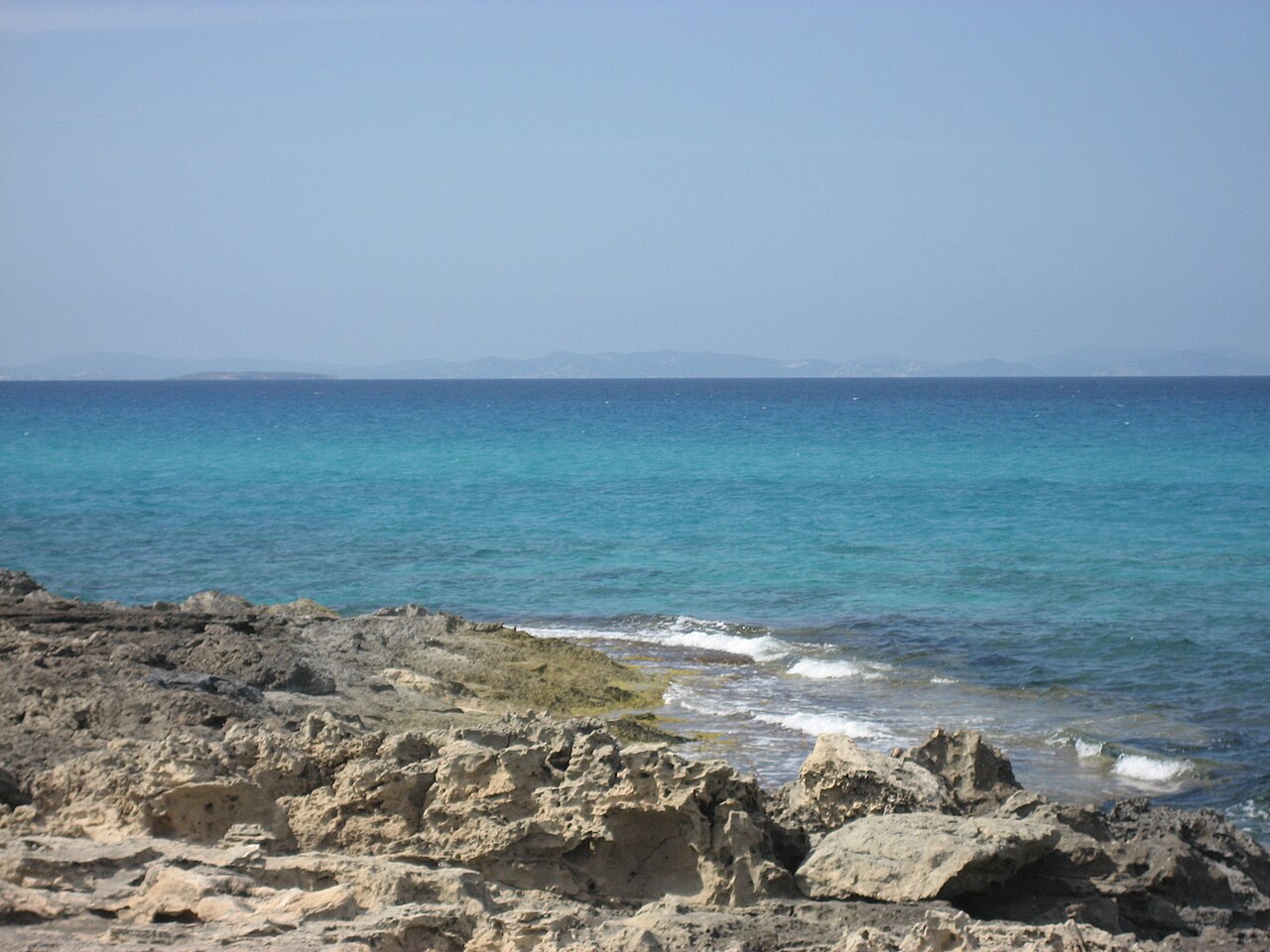 Rotsplaat aan de waterlijn bij Ses Platgetes met helder turquoise water en de heuvels van Ibiza aan de horizon