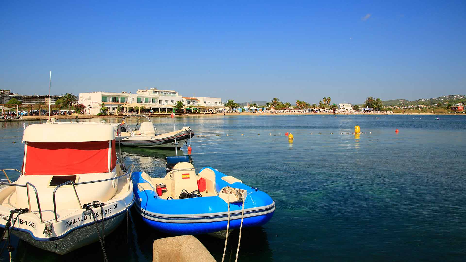 Twee bootjes afgemeerd aan een steigertje in de baai van Talamanca, met achter het ondiepe water de badlijn met boeien, het zandstrand en de witte gebouwen erachter