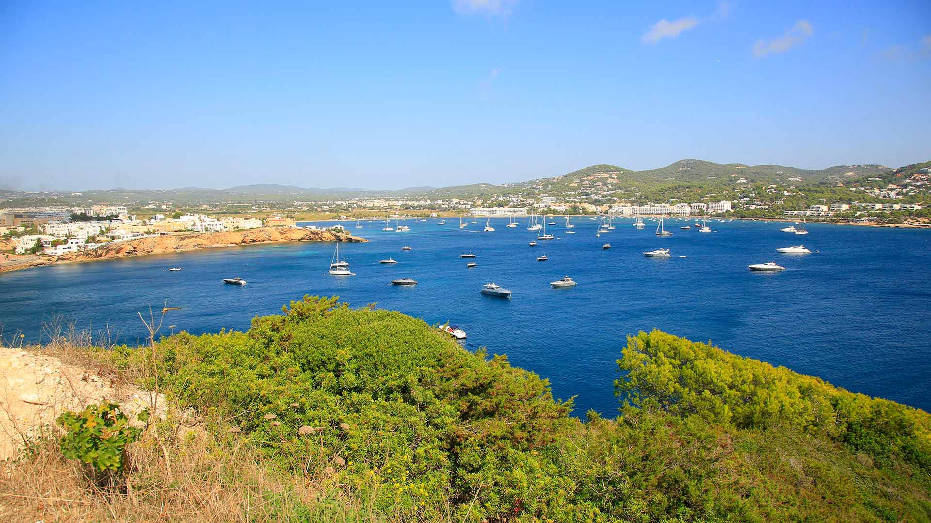 De hele baai van Talamanca van bovenaf gezien, met tientallen zeilboten en motorjachten voor anker en de bebouwing en heuvels rond de baai